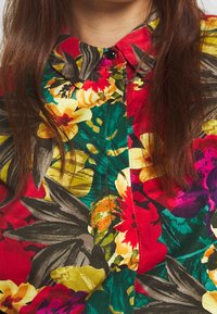 Chemise fleurie à base rouge, présentant de grandes fleurs jaunes, roses et vertes. Col boutonné et tissu brillant avec des motifs de feuilles foncées.