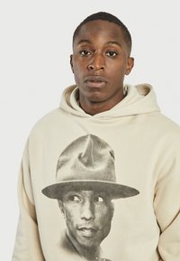Beige hættebluse med et trykt sort-hvidt portræt af en person iført en hat, med en blød struktur og enkel design.