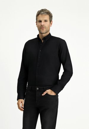 Camisa negra de manga larga con botón y cuello clásico. Tejido suave, diseño ajustado y brillo sutil. Combinada con jeans de mezclilla oscuros.
