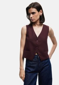 Veste sans manches bordeaux avec décolleté en V, deux boutons et poches latérales, associée à un jean bleu foncé, présentée sur un fond blanc.