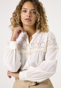 Weißes Blusenshirt mit Broderie-Anglaise-Verzierungen, langen, luftigen Ärmeln und lockerer Passform. Weicher, strukturierter Stoff mit einem zarten Muster.