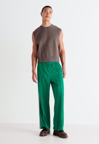 Pull sans manches marron en tissu doux ; pantalon de sport vert avec des bandes latérales ; le mannequin se tient contre un fond blanc uni.