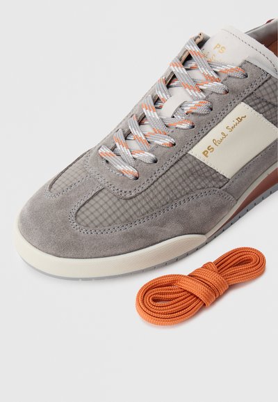 Zapatilla deportiva de ante gris con detalles de malla gris, suela blanca y cordones con detalles en naranja. Incluye un cordón adicional color naranja.