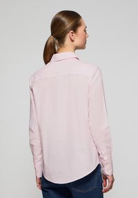 Light pink long-sleeve shirt with a pointed collar, back yoke seam detail, and rounded hemline. Buttoned cuffs display subtle stitching.