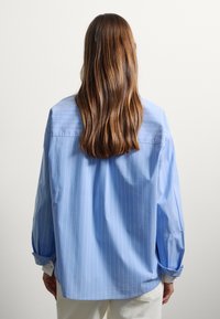 Persona con cabello largo castaño que lleva una camisa de rayas azul claro y pantalones blancos, vista desde atrás contra un fondo sencillo.