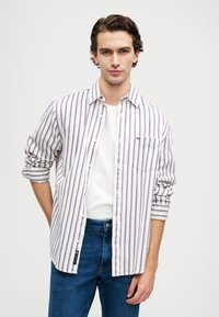 Ριγέ πουκάμισο σε λευκό χρώμα με μωβ και κόκκινες ρίγες, με μακριά μανίκια που είναι σηκωμένα, μία τσέπη στο στήθος, συνδυασμένο με μπλε τζιν.