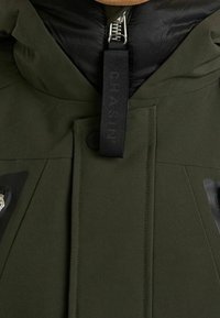 Kurtka w oliwkowym zielonym kolorze z pikowaną czarną podszewką, zamkiem błyskawicznym, widocznym przednim paskiem z wytłoczonym napisem oraz dwoma bocznymi kieszeniami na zamek.