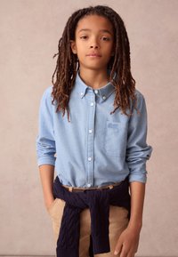 Vaaleansininen nappipaita, jossa on tasku, yhdistetty tummansiniseen neuletakkiin, joka on sidottu vyötärölle, ja beigeihin housuihin.