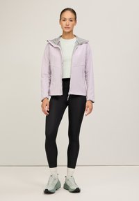 Hellviolette Sportjacke mit grauem Kapuzenrand, Reißverschluss vorne, zwei Seitentaschen, kombiniert mit schwarzen Leggings und grünen Turnschuhen. Glattes Material, figurbetontes Design.
