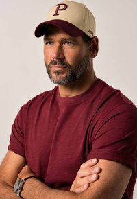 Beige cap with maroon bill, features a large 'P' logo. Maroon T-shirt with short sleeves. Silver watch on wrist, arms crossed.