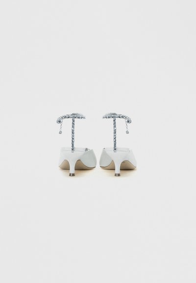 Par de sapatos brancos de salto alto com tiras finas de cristal ao redor do tornozelo, vistos por trás sobre um fundo claro e simples.