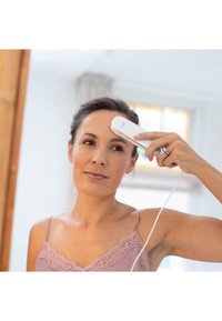 Portable facial device, white plastic with sleek design, held to forehead; connected by cord; user in soft pink attire, natural lighting.