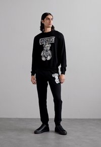 Sudadera negra con el gráfico de "Moschino" y un oso de peluche; combinada con pantalones negros que presentan un diseño similar de oso de peluche. Llevado con zapatillas negras.