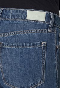 Jeans de ganga azuis com um efeito desbotado, apresentando costuras laranja, bolsos traseiros e um rótulo de couro azul claro perto da cintura.
