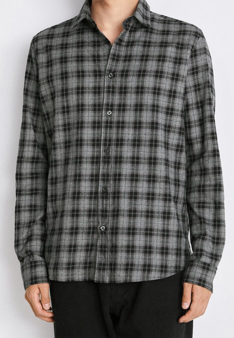 Camisa de botón en patrón de cuadros gris y negro, fabricada en una tela suave. Cuenta con un cuello, mangas largas y puños de un solo botón.