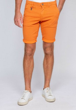 Homme portant un short orange vif retroussé et des baskets blanches, debout les mains dans les poches devant un fond uni.