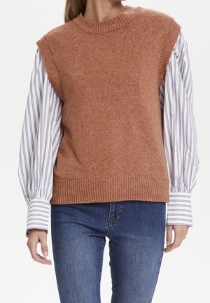 Frau in einem braunen ärmellosen Sweater-Vest über einem weißen Langarmshirt mit blauen Streifen und blauen Jeans, die mit entspannten Armen dasteht.