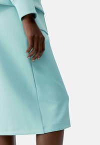 Egy közeli kép egy kézről, amely egy világoskék, sima textúrájú ruhán pihen, amelynek enyhe szélesedése és varrás részletei vannak az alján.