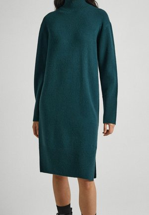 Femme portant une robe-pull à col roulé vert foncé à manches longues avec poignets et ourlet côtelés, debout devant un fond uni.