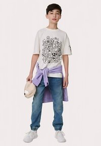 Wit T-shirt met grafische print, lichtpaarse sweatshirt om de taille gestrikt, blauwe spijkerbroek, witte sneakers en beige pet.
