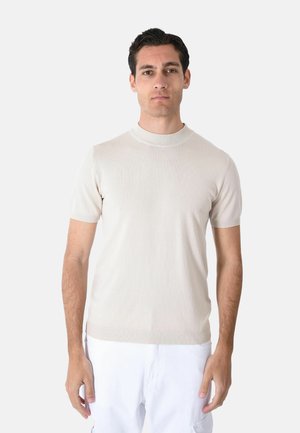 Uomo con capelli scuri che indossa un maglione beige semplice a maniche corte e pantaloni bianchi, in piedi davanti a uno sfondo chiaro, volto verso avanti.