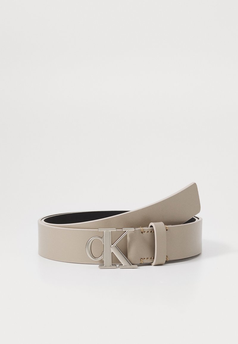 Beige műbőr öv ezüst színű "CK" logós csattal. Simább textúrájú, lapos kialakítású, és két kis arany szegecs található a csatt közelében.