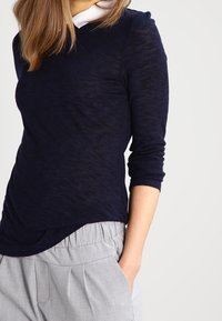 Maglia a maniche lunghe blu marino con un motivo testurizzato e scollo rotondo, abbinata a pantaloni grigi chiari plissettati, mani nelle tasche.