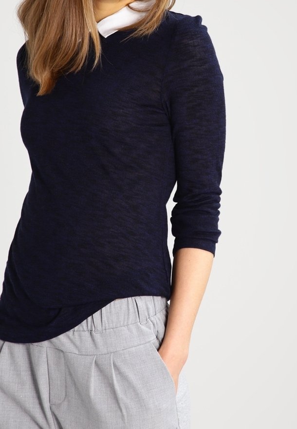 Maglia a maniche lunghe blu marino con un motivo testurizzato e scollo rotondo, abbinata a pantaloni grigi chiari plissettati, mani nelle tasche.