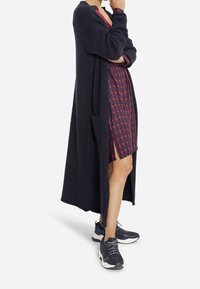Dlouhý tmavě modrý kardigan s texturovanou látkou, otevřeným předním dílem a bočními kapsami, nošený přes vzorované šaty a doplněný o tmavé tenisky.