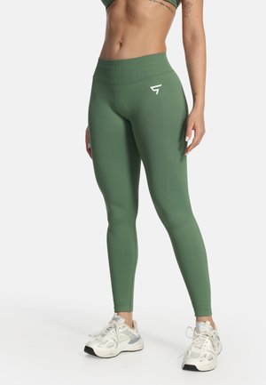 Frau trägt grüne, hochgeschnittene Leggings mit einem Logo an der Hüfte, kombiniert mit weißen Sportschuhen, und steht vor einem weißen Hintergrund.