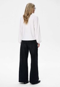 Weißes Langarmoberteil mit rundem Kragen, kombiniert mit hochbundigen, schwarzen Weitjeans, die flache Nähte und zwei Gesäßtaschen aufweisen.