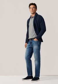 Hombre vistiendo un cárdigan azul marino, camiseta polo gris, jeans azules y zapatillas negras, de pie con una mano en el bolsillo frente a un fondo liso.