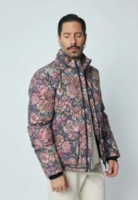 Veste matelassée florale en gris foncé avec des motifs de fleurs roses, vertes et crème. Elle possède un col haut, une fermeture éclair et des poches latérales.