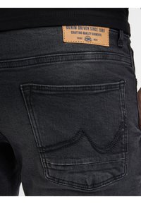 Black denim jeans featuring contrasting stitching, a leather patch labeled "DENIM DRIVEN SINCE 1989," and back pockets with decorative stitching.