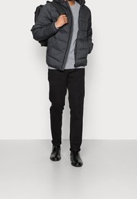 Dunjakke i mørkegrå, tekstureret stof, lynlåslukning foran; grå trøje under. Sorte bukser og polerede sorte ankelstøvler fuldender looket.