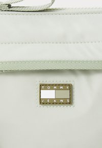 Sac Tommy Jeans vert clair en matériau lisse, avec des accents cousus et une plaque de logo en métal rectangulaire avec du texte.