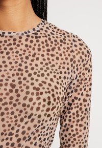 Läpinäkyvä yläosa, jossa on pyöreä pääntie, beige tausta ja tummanruskeita ja mustia laikkuja kauttaaltaan. Pitkät hihat.