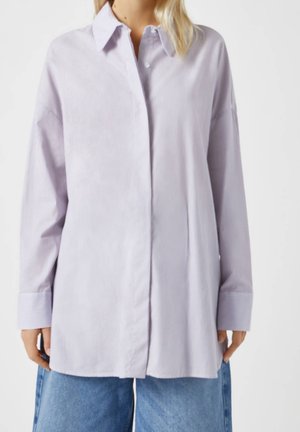 Kvinde iført en løs, lyse lilla stribet skjorte med knapper, lange ærmer og blå jeans mod en ensfarvet baggrund.