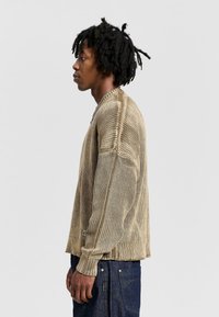 Beige knitted sweater with ribbed texture, round neckline, and long sleeves. Worn with dark denim pants, viewed from the side.