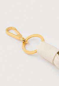 Breloc metalic auriu și clemă atașată la o curea texturată albă din piele, cu logo-ul Coccinelle pe inel.