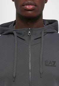 Gray zip-up hoodie with a drawstring hood, featuring a subtle logo on the chest and textured fabric for a comfortable fit.