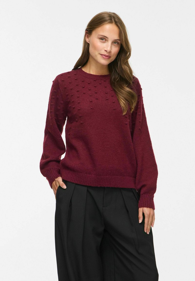 Femme aux longs cheveux bruns portant un pull bordeaux texturé et un pantalon noir ample, debout avec une main dans la poche, sur fond blanc.