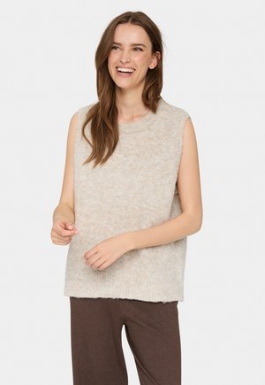 Beige, bolyhos, ujjatlan felső kerek nyakkivágással, puha anyagból készült. Lazán illeszkedő fazon, egyenes szegéllyel, barna nadrággal viselve.
