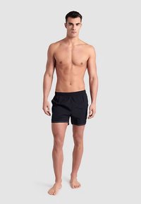 Zwarte zwembroek met een elastische tailleband, verstelbare trekkoord en witte sierstiksels. Model staat blootsvoets in een neutrale houding.