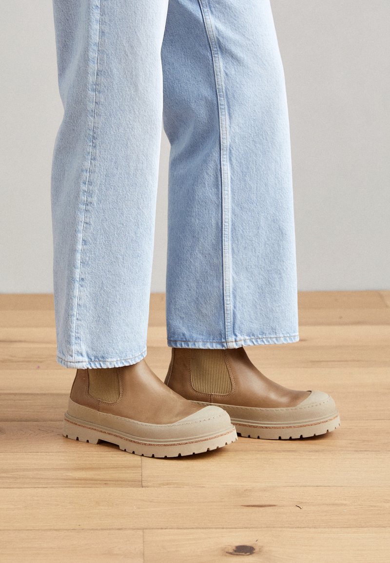 Stivali Chelsea in pelle marrone con pannelli elastici sui lati e suole in gomma beige con finitura texture, indossati con pantaloni in denim azzurro chiaro.