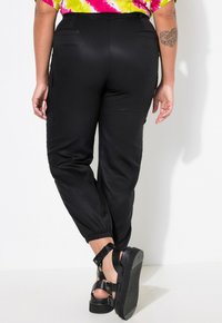 Schwarze Cargo-Hosen mit elastischen Bündchen, ausgestattet mit seitlichen Taschen und einer glatten Textur. Kombiniert mit klobigen schwarzen Sandalen mit gerippter Sohle.