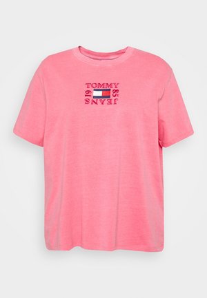 Roze T-shirt met korte mouwen, ronde hals en rode geborduurde tekst "Tommy Jeans 85 19" die een rechthoekig rood, wit en blauw logo op de borst omlijst.