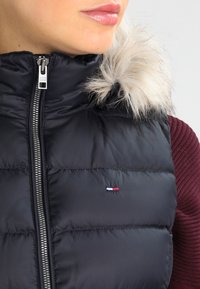 Svart pufferväst med huva i konstpäls. Har en silverdragkedja och en liten logotyp i rött, vitt och blålängst fram.