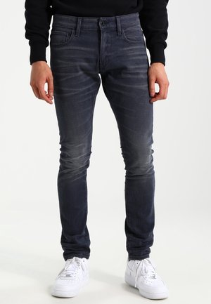 Jeans de mezclilla oscuros de corte ajustado con un ligero desgaste, cinco bolsillos y cremallera, combinados con zapatillas blancas.
