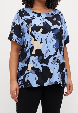 Kobieta ubrana w krótką, luźną bluzkę z abstrakcyjnym wzorem w kolorach niebieskim, czarnym i beżowym oraz czarne spodnie, stojąca na białym tle.